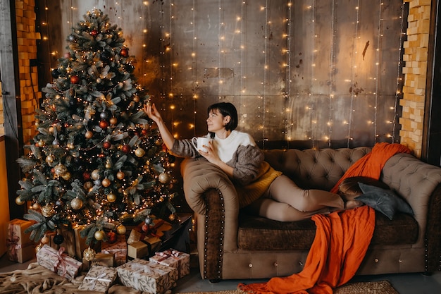 Mujer sentada en el sofá con una taza de café o té junto a un árbol de Navidad en casa