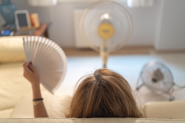 Mujer sentada en un sofá con varios ventiladores dándole aire y un ventilador en la mano altas temperaturas veraniegas