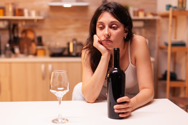 Mujer sentada sola con una botella de vino tinto en la cocina debido a la expresión. Persona infeliz que sufre de migraña, depresión, enfermedad y ansiedad sintiéndose agotada.