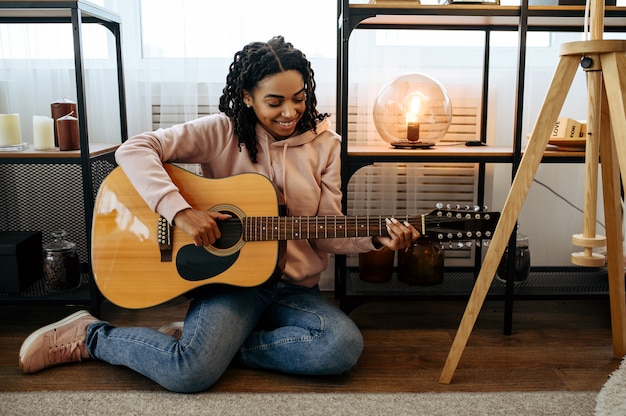 Mujer sentada en el suelo y tocar la guitarra en casa, vista de cerca. Señora bonita con instrumento musical relajarse en la habitación, amante de la música femenina descansando