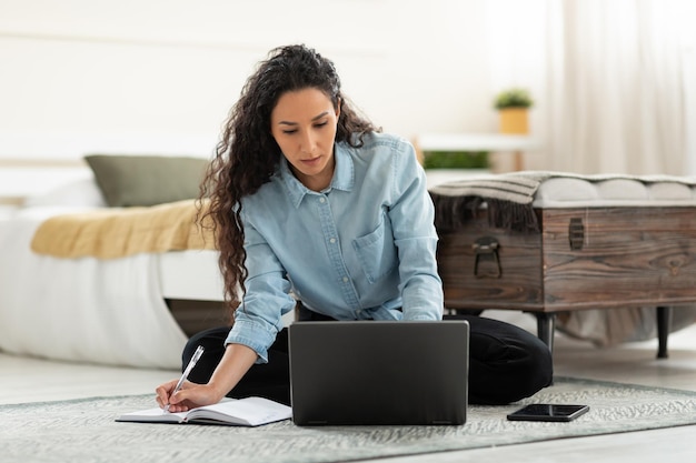 Mujer sentada en el suelo usando la computadora y escribiendo en el cuaderno