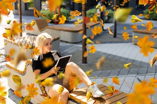 mujer sentada con una tableta en las manos cae en el parque de la ciudad en un día cálido. Hojas doradas de otoño.