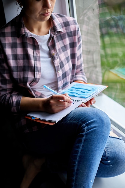 Mujer sentada en la ventana, escribiendo en un plan de cuaderno, para hacer la lista después del final de la cuarentena, finalización del coronovirus