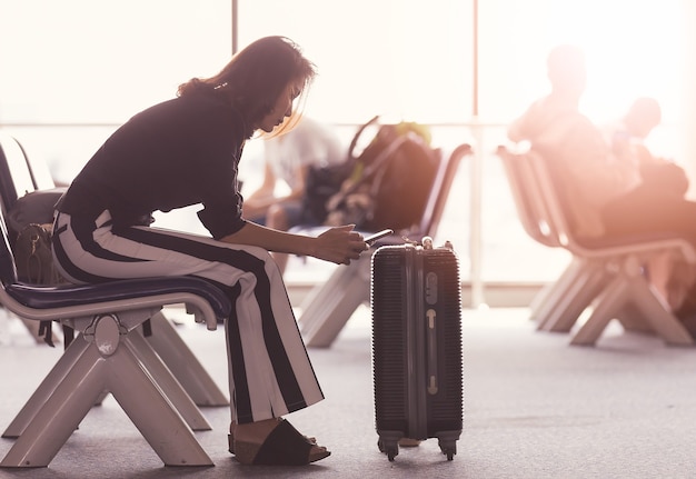 Mujer sentada en el vestíbulo del aeropuerto con smartphone y mirando la pantalla mientras espera el tránsito. La luz del sol brilla desde el exterior.