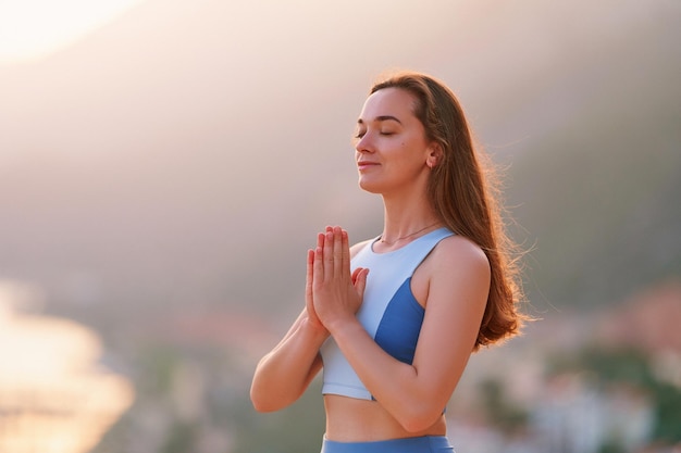 Una mujer serena, esperanzada e inspirada, medita con los ojos cerrados y se para sola con las manos rezando al atardecer. Alivie el estrés y mente limpia y sana.