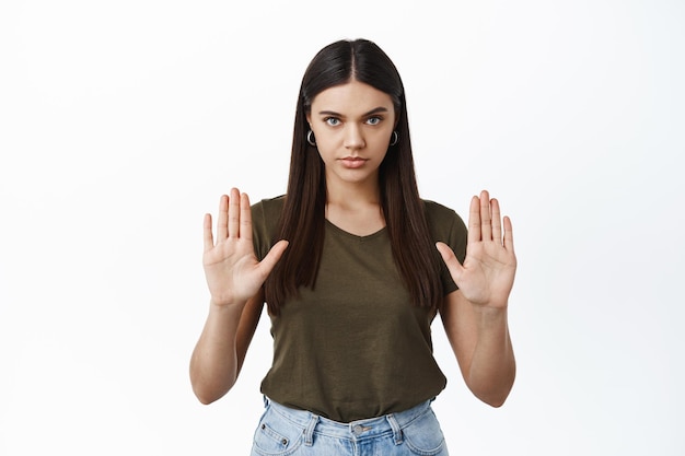 Foto una mujer seria bloquea su oferta, muestra la señal de alto, no hace ningún gesto y mira al frente decidida a negarse, de pie sobre una pared blanca