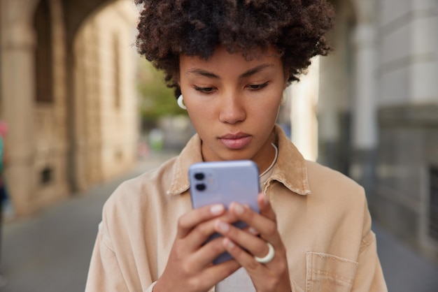 Una mujer seria de cabello rizado envía mensajes de texto a través de búsquedas de teléfonos inteligentes en las calles de la ciudad busca en la dirección correcta usa la aplicación usa una chaqueta informal Gente tecnologías modernas y concepto de estilo de vida