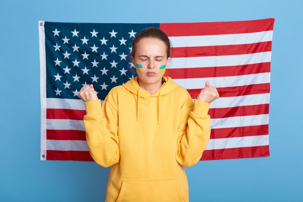 Mujer seria y concentrada con capucha amarilla con bandera ucraniana en las mejillas posando aislada sobre fondo azul con bandera estadounidense contra ella de pie con los puños cerrados