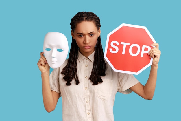 Mujer seria con dreadlocks negros con máscara blanca y señal de stop roja, mirando a la cámara, no cambia de personalidad, usando camisa blanca. Disparo de estudio interior aislado sobre fondo azul.