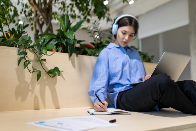 Foto mujer seria y enfocada con auriculares sentada en un espacio de trabajo haciendo notas en el cuaderno