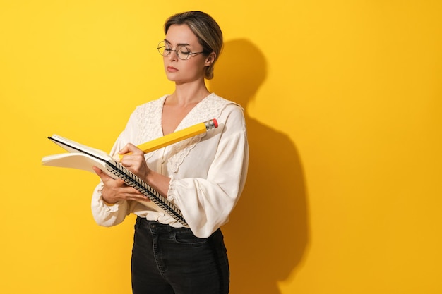 Mujer seria con gafas sosteniendo un lápiz grande y un cuaderno sobre fondo amarillo