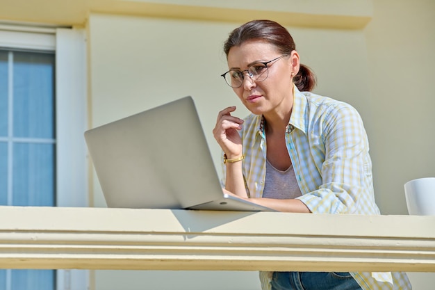 Mujer seria de mediana edad con laptop en el balcón exterior de la casa