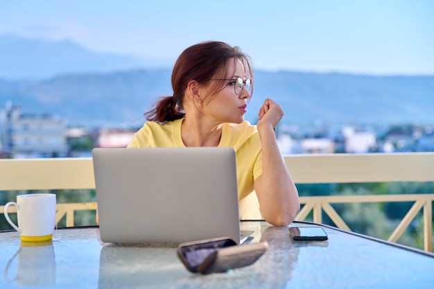 Mujer seria de mediana edad con portátil en la mesa en el balcón al aire libre de la casa