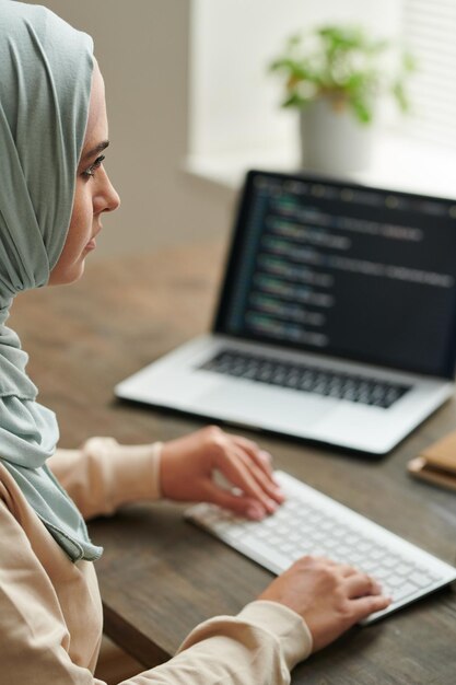 Mujer seria del medio oriente usando hiyab sentada en el escritorio trabajando en un disparo vertical de código de computadora