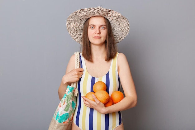 Mujer seria y segura de sí misma con traje de baño a rayas de colores de pie con bolsa y naranjas aisladas sobre fondo gris mirando a la cámara con expresión tranquila