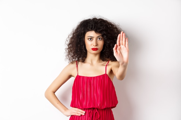 Mujer seria y segura en vestido rojo y maquillaje estirar la mano, decir que se detenga, prohibir y prohibir algo, de pie sobre fondo blanco.