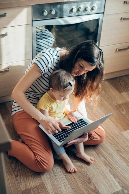 Una mujer seria sentada en el piso de la cocina con una laptop moderna y viendo a su hija pequeña tocar el teclado