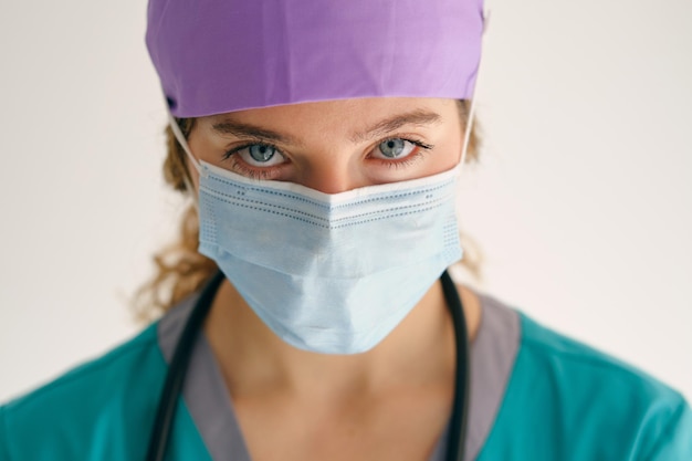 Mujer seria con uniforme médico y máscara protectora mirando a la cámara con preocupación mientras está de pie en un fondo beige