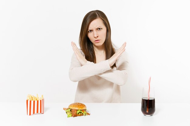 Mujer severa muestra gesto de parada en la mesa con cola de hamburguesa papas fritas en botella de vidrio aislado sobre fondo blanco. Nutrición adecuada o comida rápida clásica estadounidense. Área de publicidad con espacio de copia.