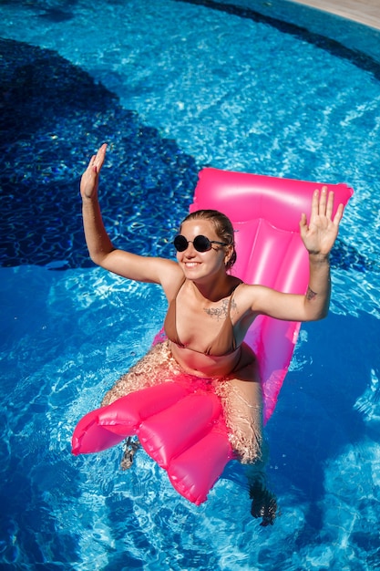 Mujer sexy en gafas de sol descansando y tomando el sol en un colchón rosa en la piscina. Mujer joven en traje de baño bikini beige flotando sobre un colchón inflable rosa