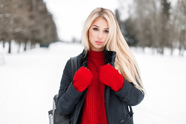 Mujer sexy hermosa joven modelo en un elegante abrigo de invierno con un suéter de punto rojo al aire libre en un día de invierno cubierto de nieve