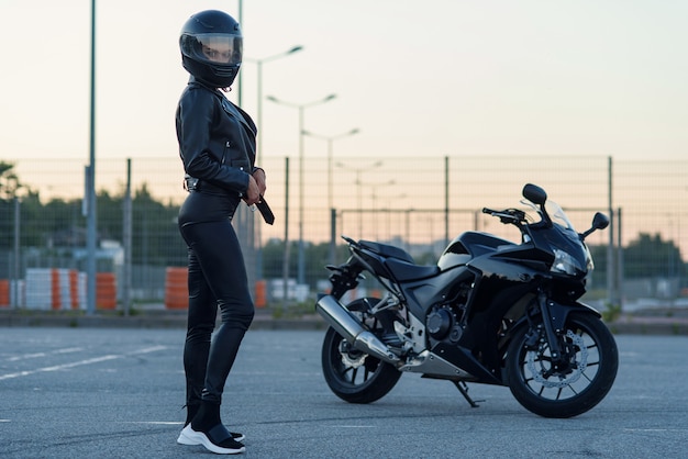 Mujer sexy motociclista en chaqueta de cuero negro y casco integral se encuentra cerca de elegante motocicleta deportiva. Aparcamiento urbano, puesta de sol en la gran ciudad. Viajar y estilo de vida activo hipster. Poder niñas.