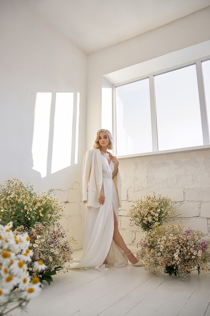 Mujer sexy romántica con una chaqueta y un vestido largo blanco está de pie cerca de la ventana en flores de manzanilla