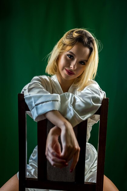 Mujer sexy en túnica blanca sentada en el estudio en una silla de madera sobre fondo liso y sonriendo