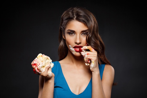 Mujer sexy en vestido azul comiendo pastel sobre pared negra