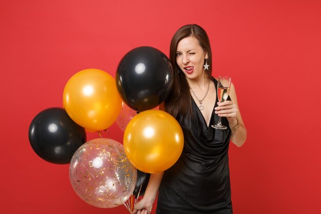 Mujer sexy en vestido negro celebrando parpadear lamiendo los labios, sosteniendo una copa de champán, globos de aire aislados sobre fondo rojo. Día de San Valentín, concepto de fiesta de vacaciones de maqueta de cumpleaños de feliz año nuevo.