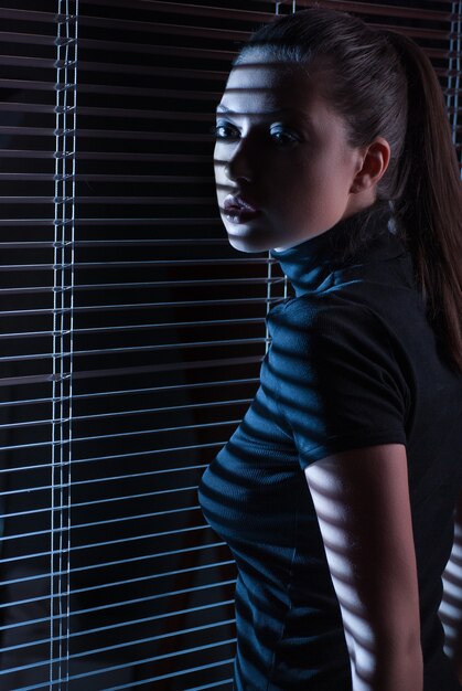 Mujer sexy en vestido negro con persianas mirando por la ventana con persianas