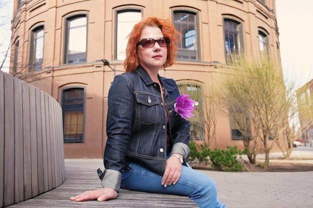 Una mujer se sienta en un banco frente a un edificio con una flor en la chaqueta.