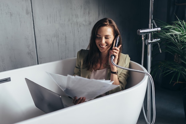 La mujer se sienta en la bañera y habla en broma en el cabezal de la ducha como si estuviera hablando por teléfono.