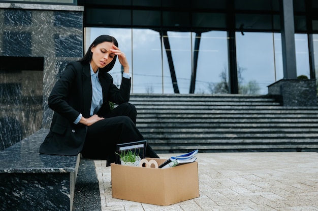 Una mujer se sienta en una caja de cartón con aspecto molesto y angustiado después de experimentar la pérdida de trabajo y el desempleo.