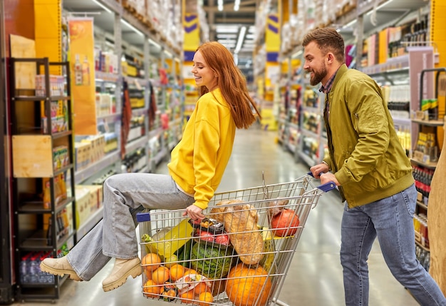 La mujer se sienta en un carro lleno de comida, el chico lleva el carro. Pareja de enamorados despreocupados divertirse durante una compra en el supermercado