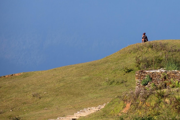 Una mujer se sienta en una colina con vista a un camino que es azul y el océano está al fondo.