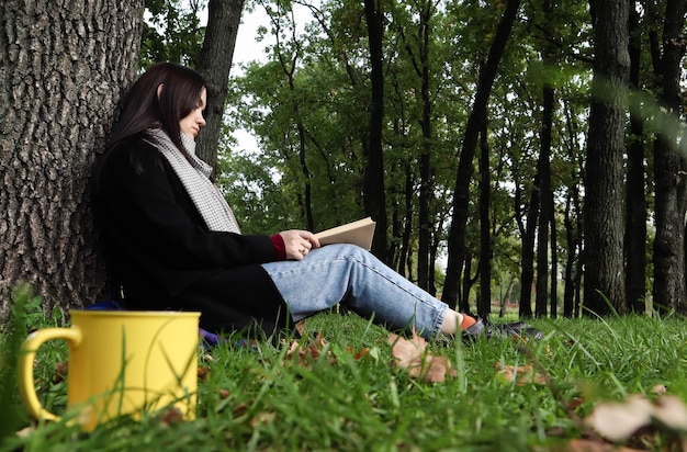 Una mujer se sienta debajo de un árbol y lee su libro favorito mientras bebe café o té de una taza amarilla en un parque de la ciudad sobre la hierba verde en un agradable día soleado. Concepto de vacaciones, educación y estudio.