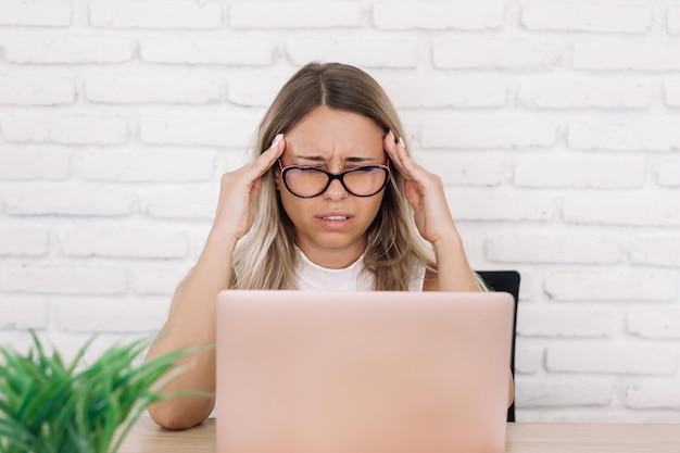 Una mujer se sienta en un escritorio con una computadora portátil con dolor de cabeza sosteniendo sus sienes con las manos