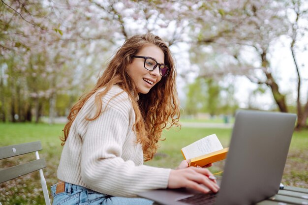 La mujer se sienta en la hierba verde cerca de los árboles en flor con una computadora portátil escribiendo en el bloc de notas el concepto de planificación