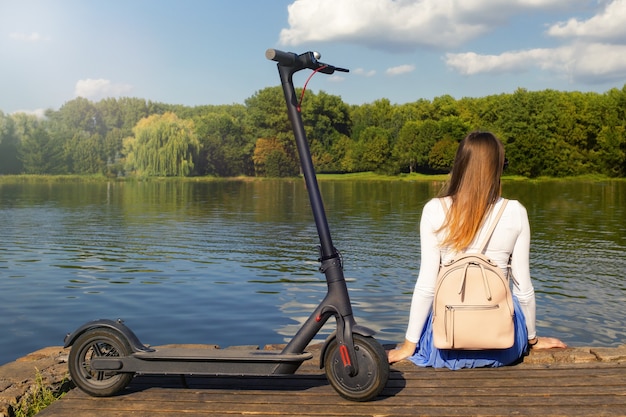 La mujer se sienta en el muelle y disfruta de la naturaleza cerca de un scooter eléctrico ecológico
