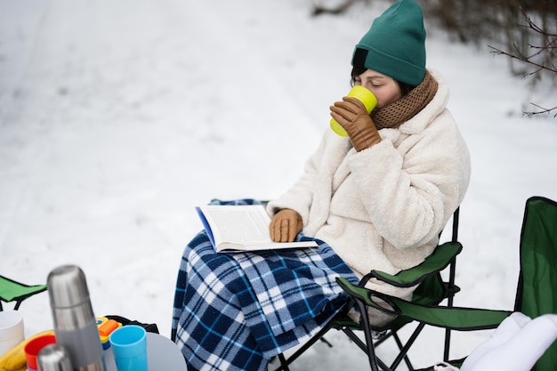 La mujer se sienta en la silla, lee el libro y bebe té en el bosque de invierno en un picnic