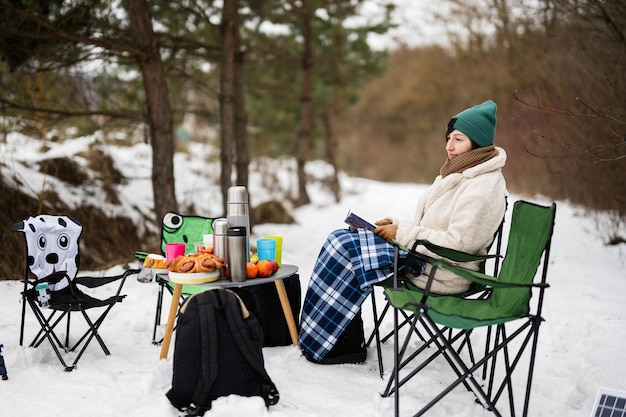 La mujer se sienta en una silla y lee un libro en el bosque de invierno en un picnic