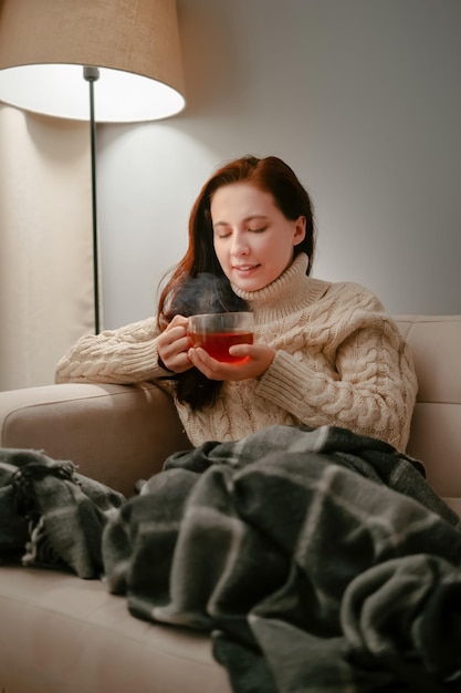 La mujer se sienta en un sofá con una taza de té curativo en una noche de invierno