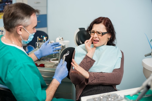 Mujer siente dolor de muelas, tocando la mejilla con la mano en la clínica dental. Dentista senior tratando de ayudar con herramienta dental - removedor