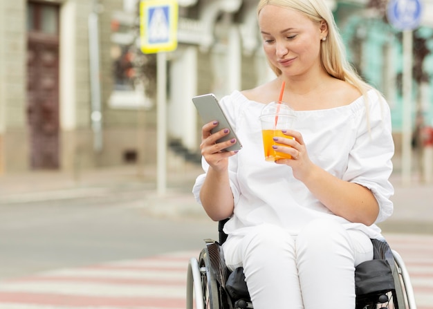 Mujer en silla de ruedas tomando una copa y usando un teléfono inteligente con espacio de copia