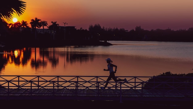 Foto mujer silueta corriendo por el puente en la orilla del lago contra la puesta de sol