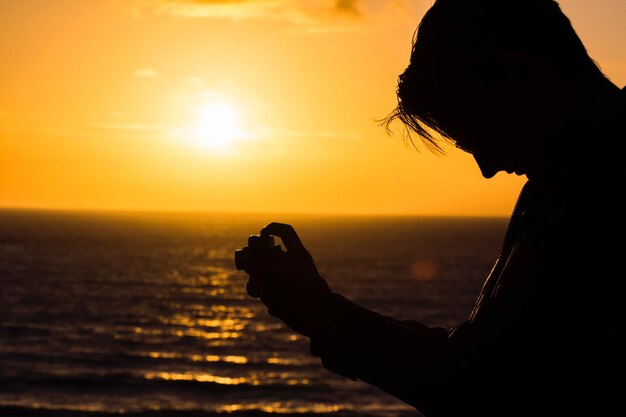 Mujer silueta fotografiando contra el mar durante la puesta de sol