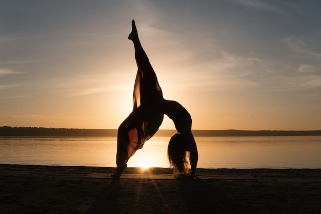 Mujer de silueta en la playa al atardecer haciendo asanas de yoga. Entrenamiento matutino de calentamiento con estiramiento natural