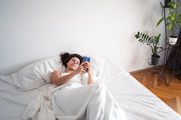 Mujer con smartphone en la cama con sábanas blancas