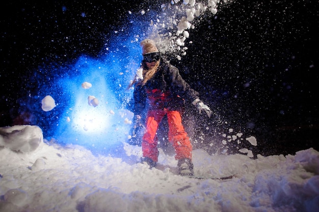 Mujer snowboarder vestida con una ropa deportiva verde de pie en la ladera de la montaña en la noche bajo la luz azul